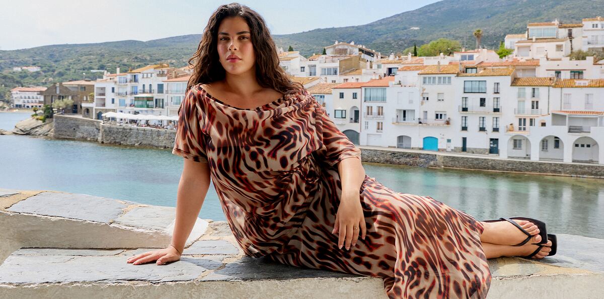 Woman reclining by a harbour in a flowing animal-print dress with a coastal village backdrop.