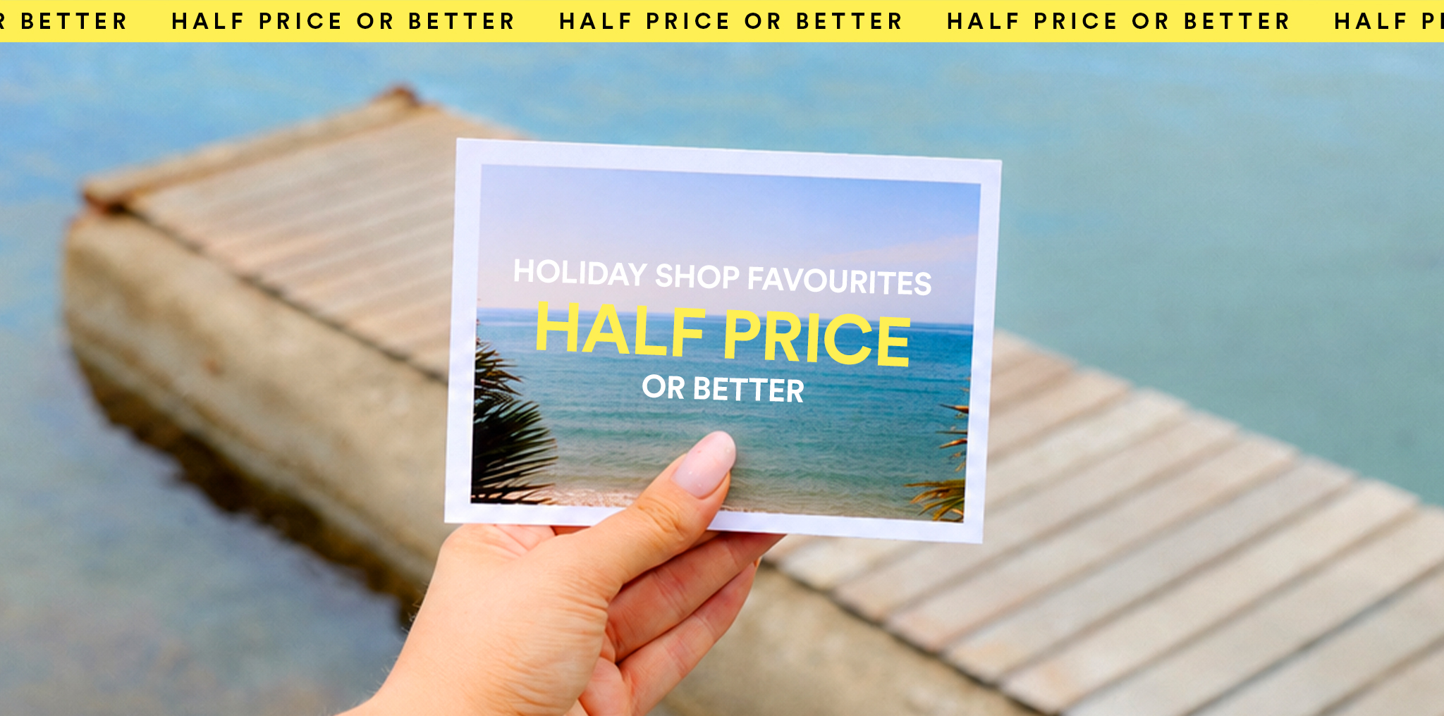 Hand holding a beach postcard with Holiday Shop Favourites Half Price or Better over a seaside dock.