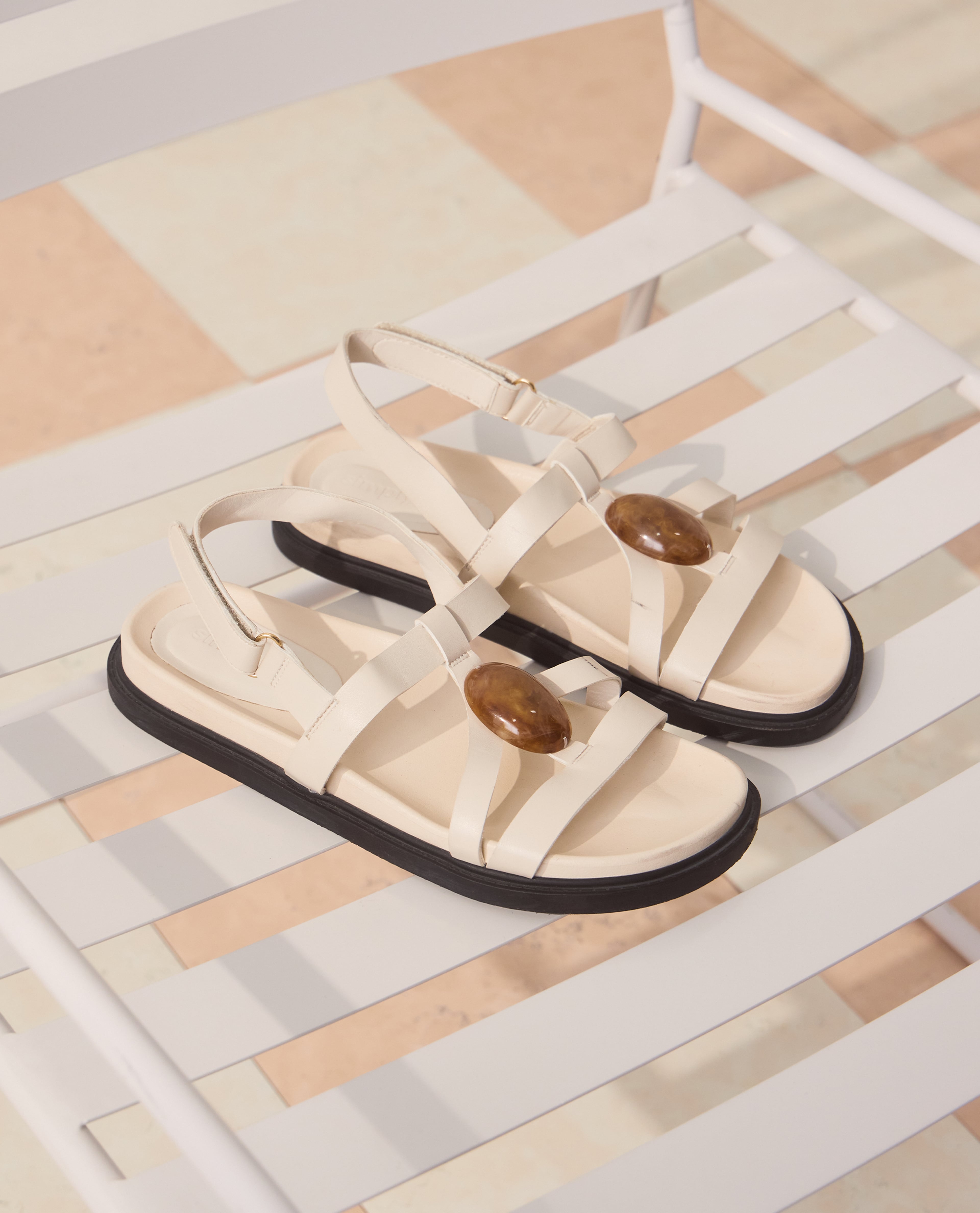 Cream strappy flat sandals with brown oval embellishments, placed on a white slatted chair in soft daylight.