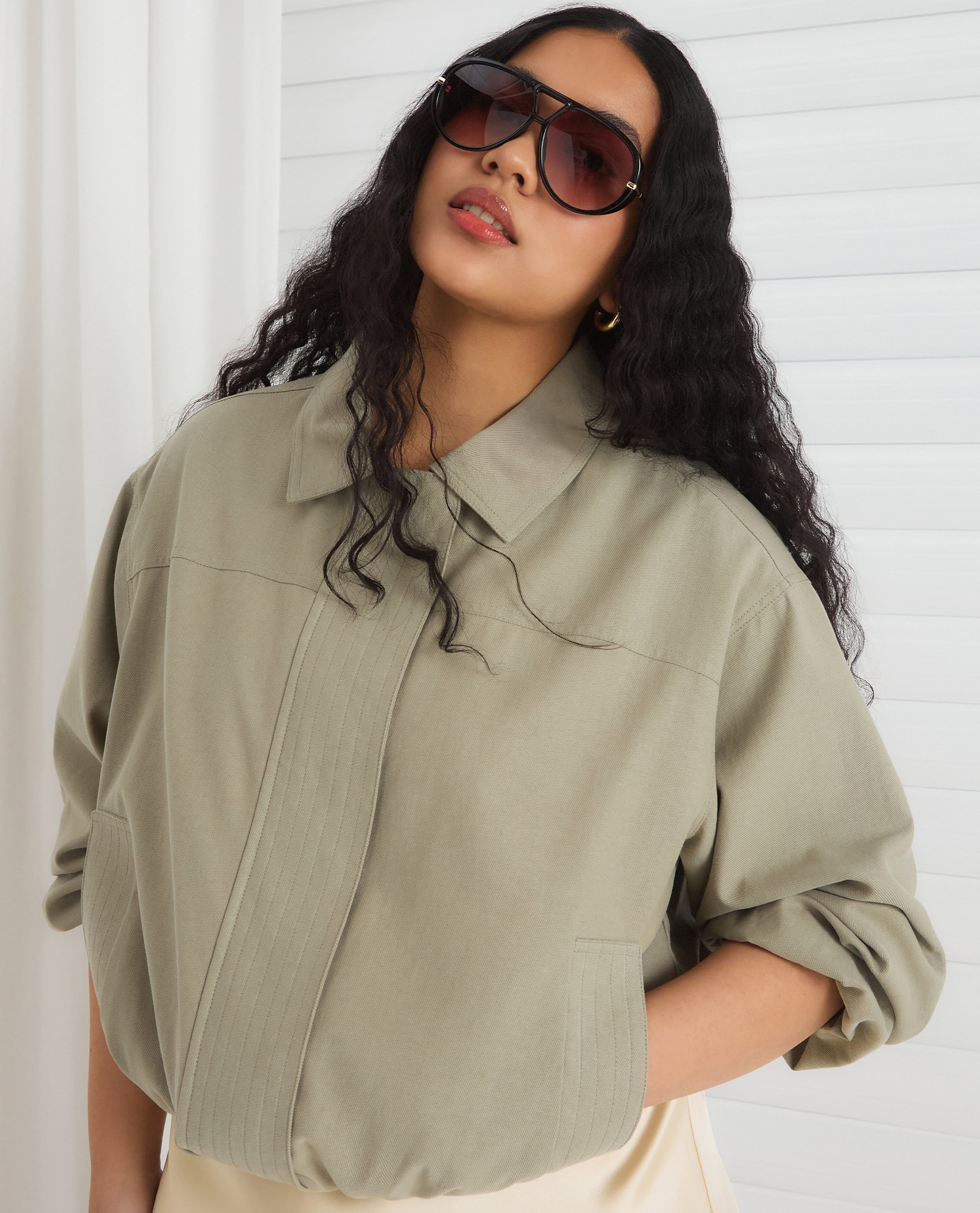 Close-up of a person wearing oversized sunglasses and a loose sage green shirt against a white backdrop.