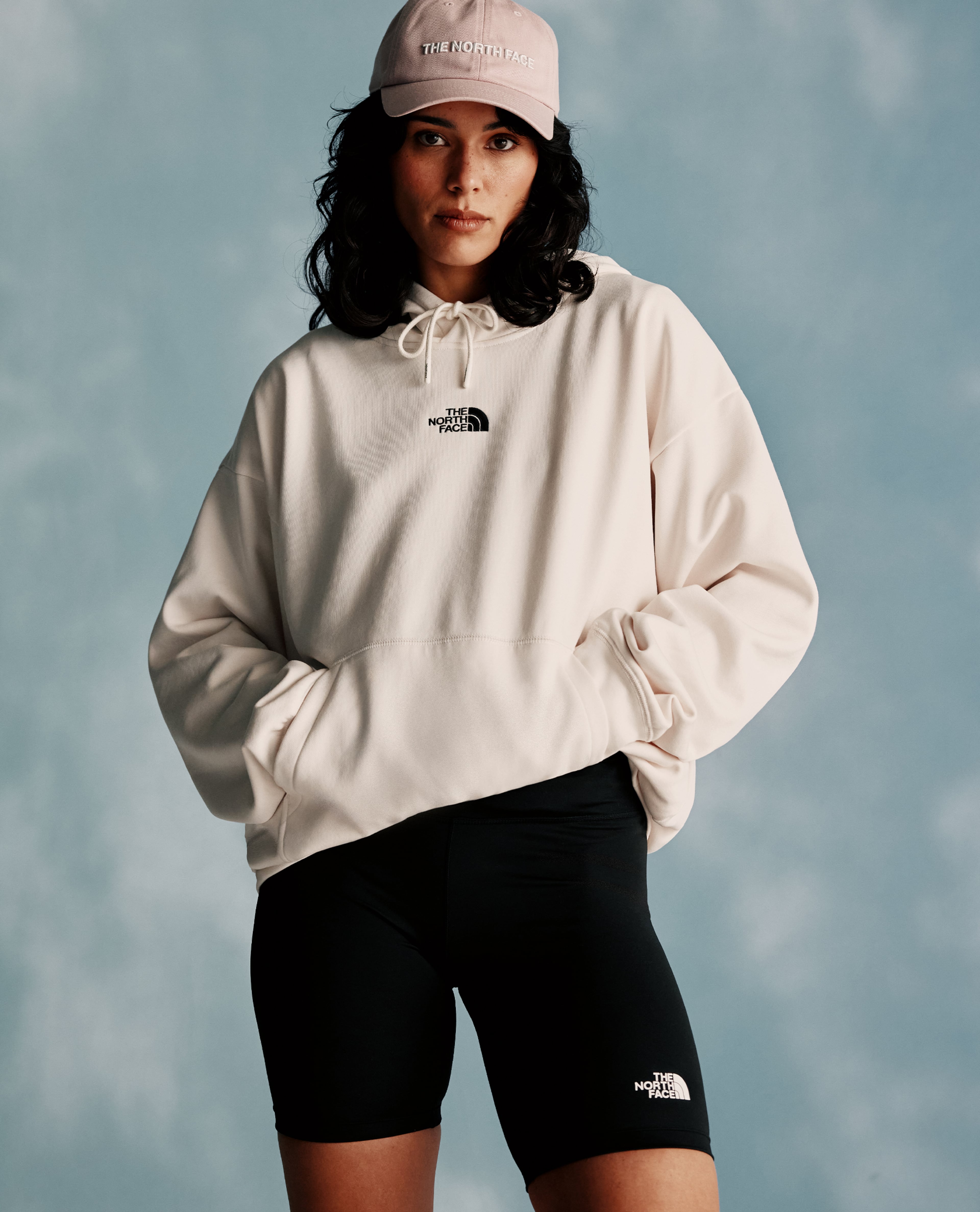 Person wearing a beige hoodie, black cycling shorts, and a logo cap against a blue studio backdrop.