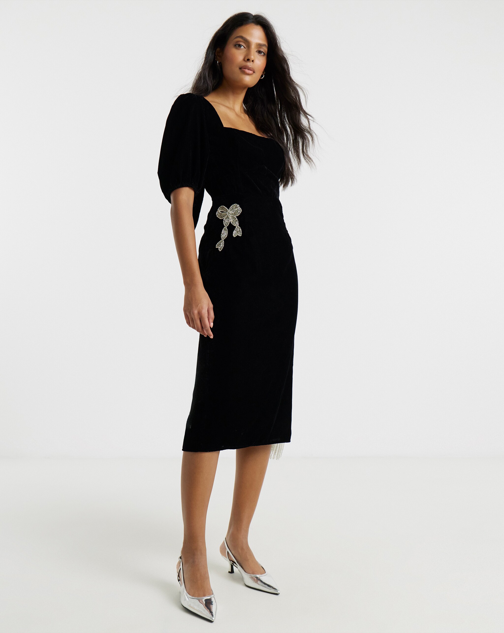 Person wearing a fitted black midi dress with puff sleeves and a bow detail at the waist, paired with metallic silver slingback heels.