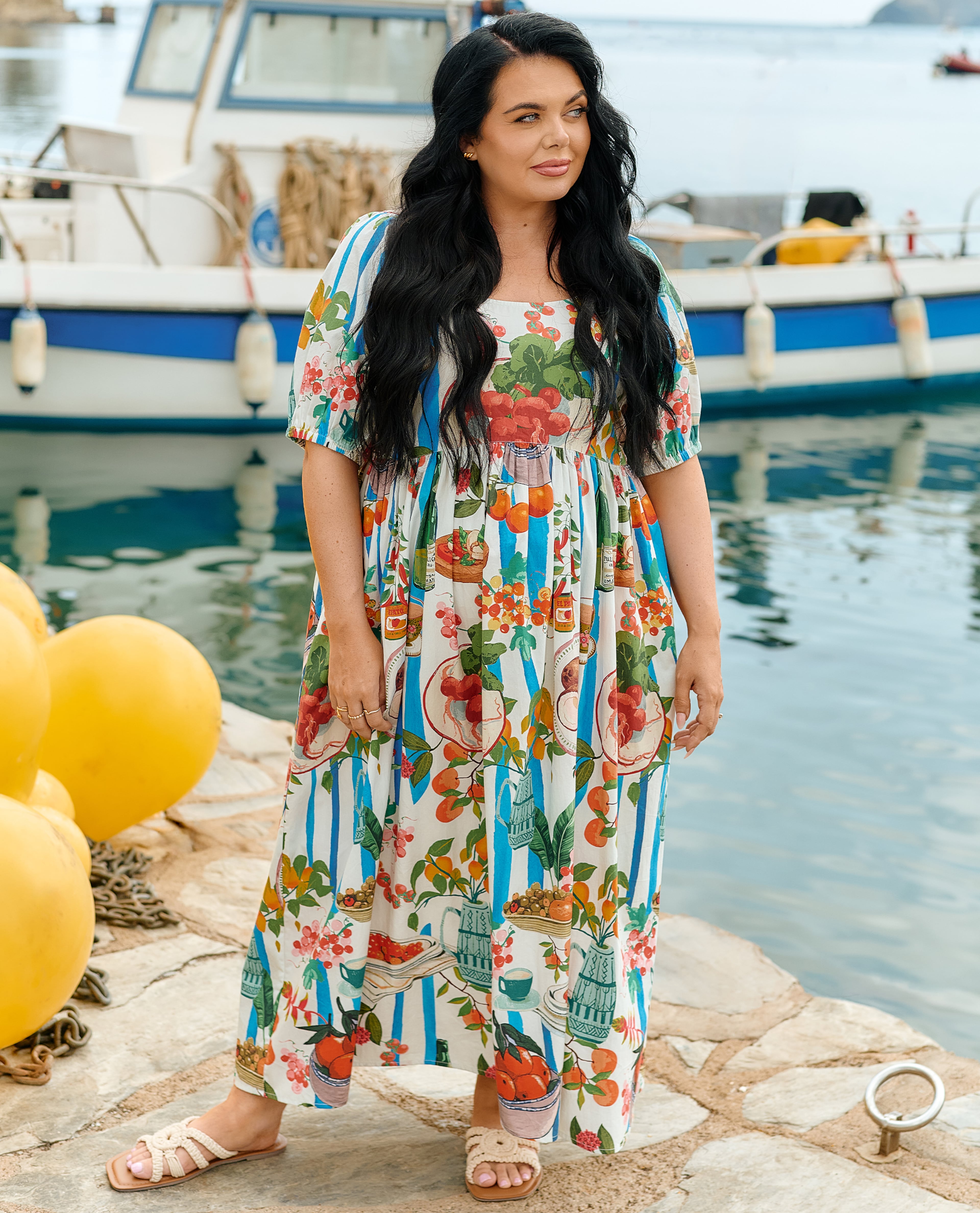 Person standing by a harbour wearing a colourful striped and fruit-print midi dress and sandals