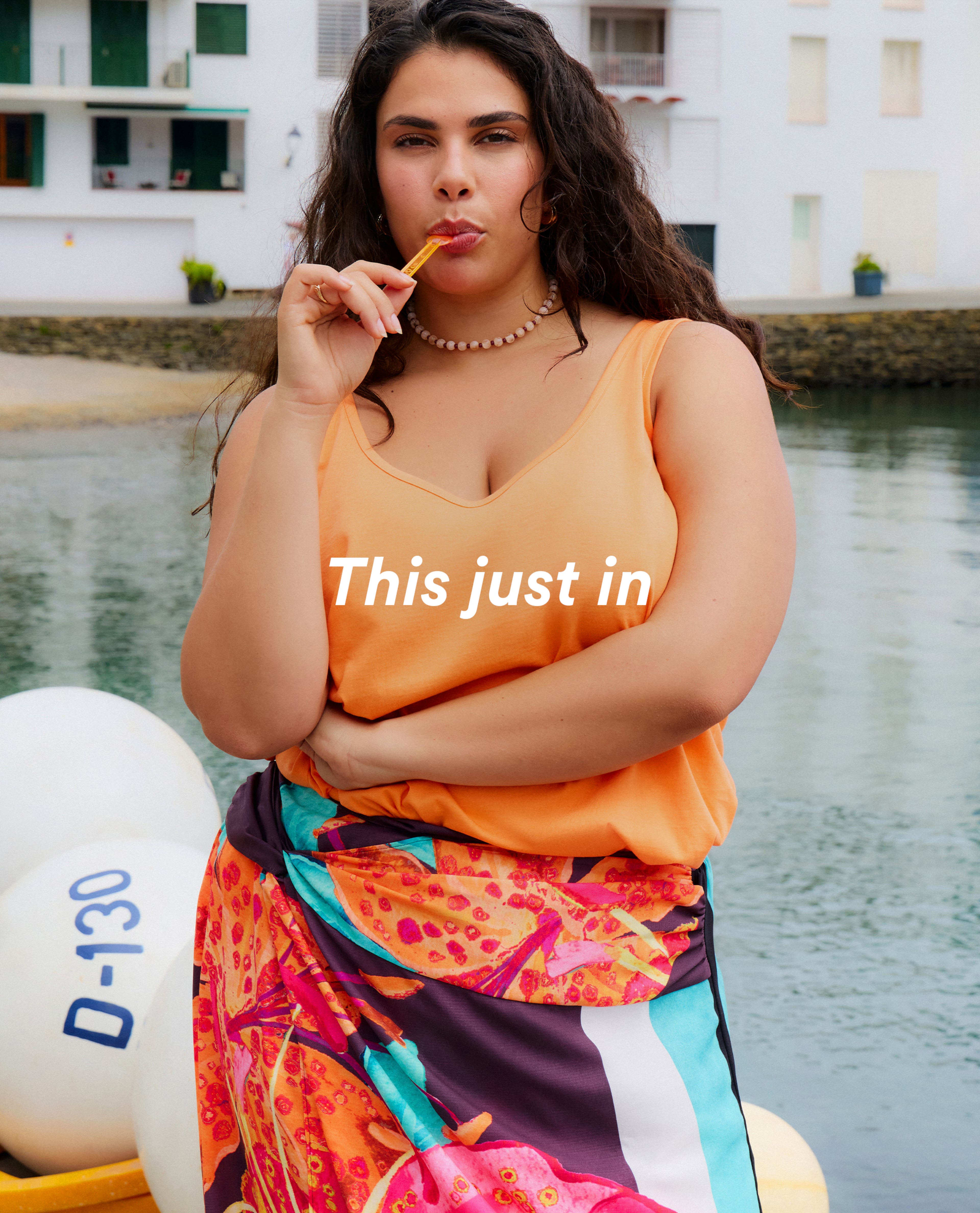 Model in an orange sleeveless top and colourful wrap skirt standing by the water.