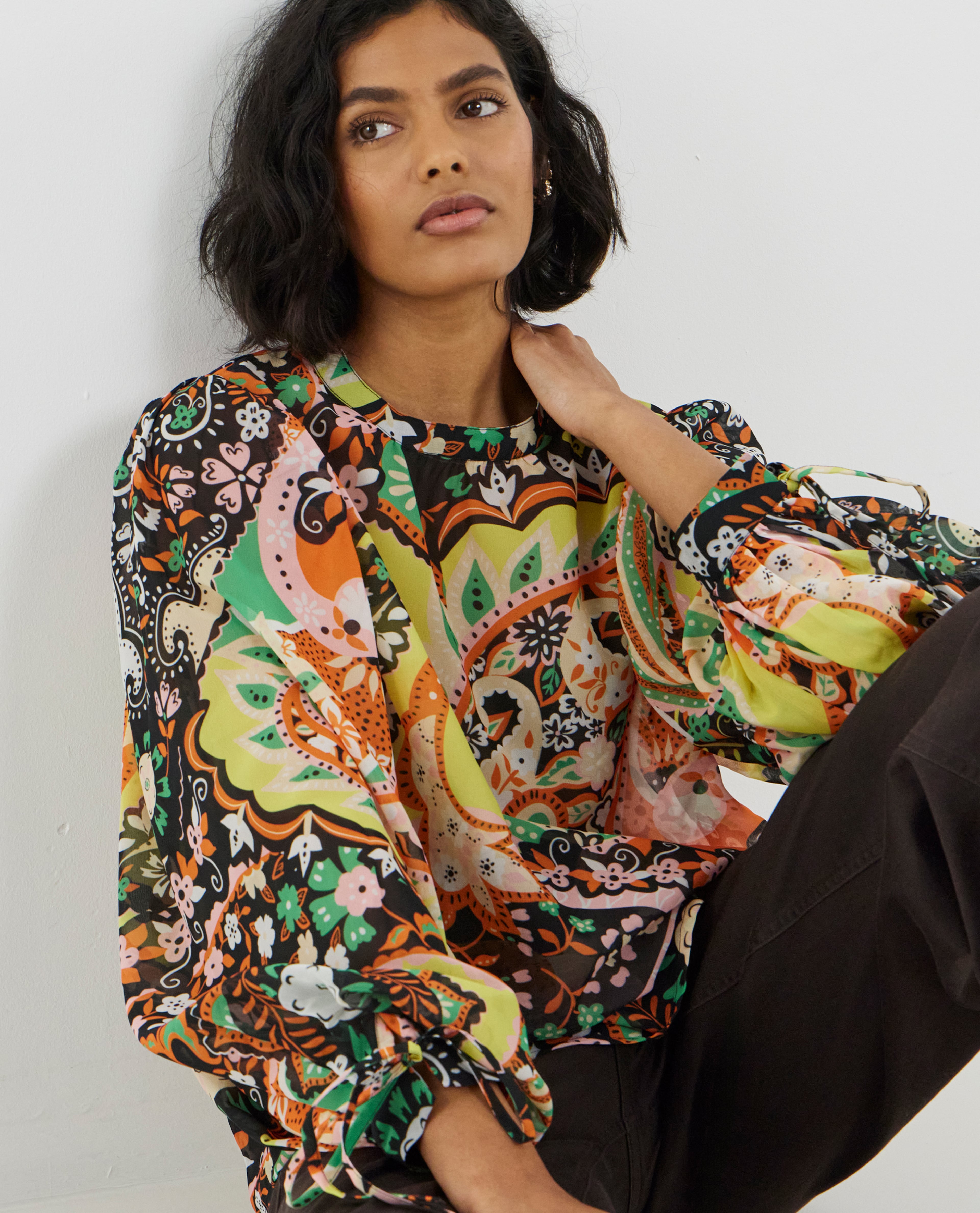 Woman wearing a colourful floral print long-sleeve blouse with black trousers, seated against a white wall.