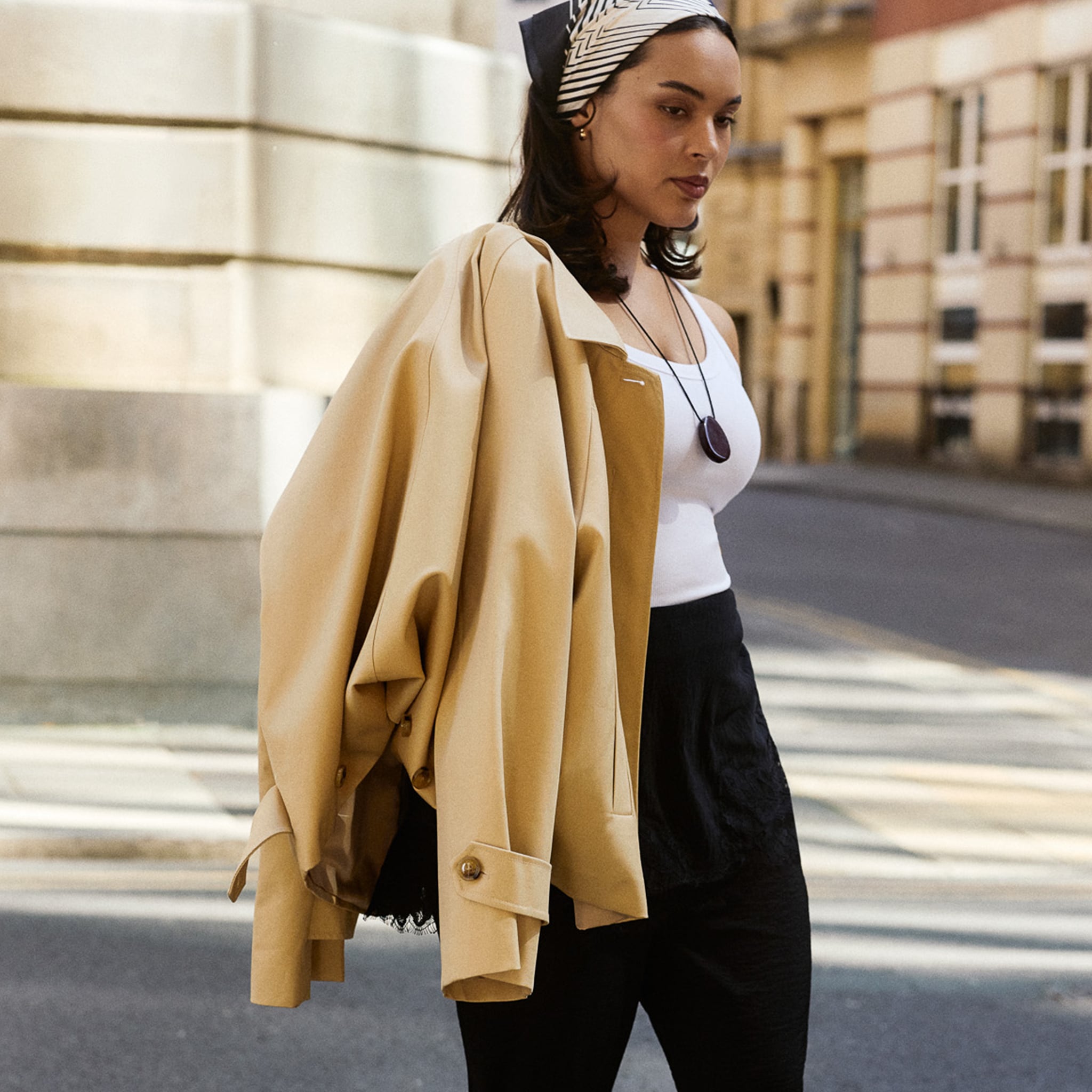 Model is wearing a head scarf, white vest top with black trousers and a camel jacket over her shoulder.