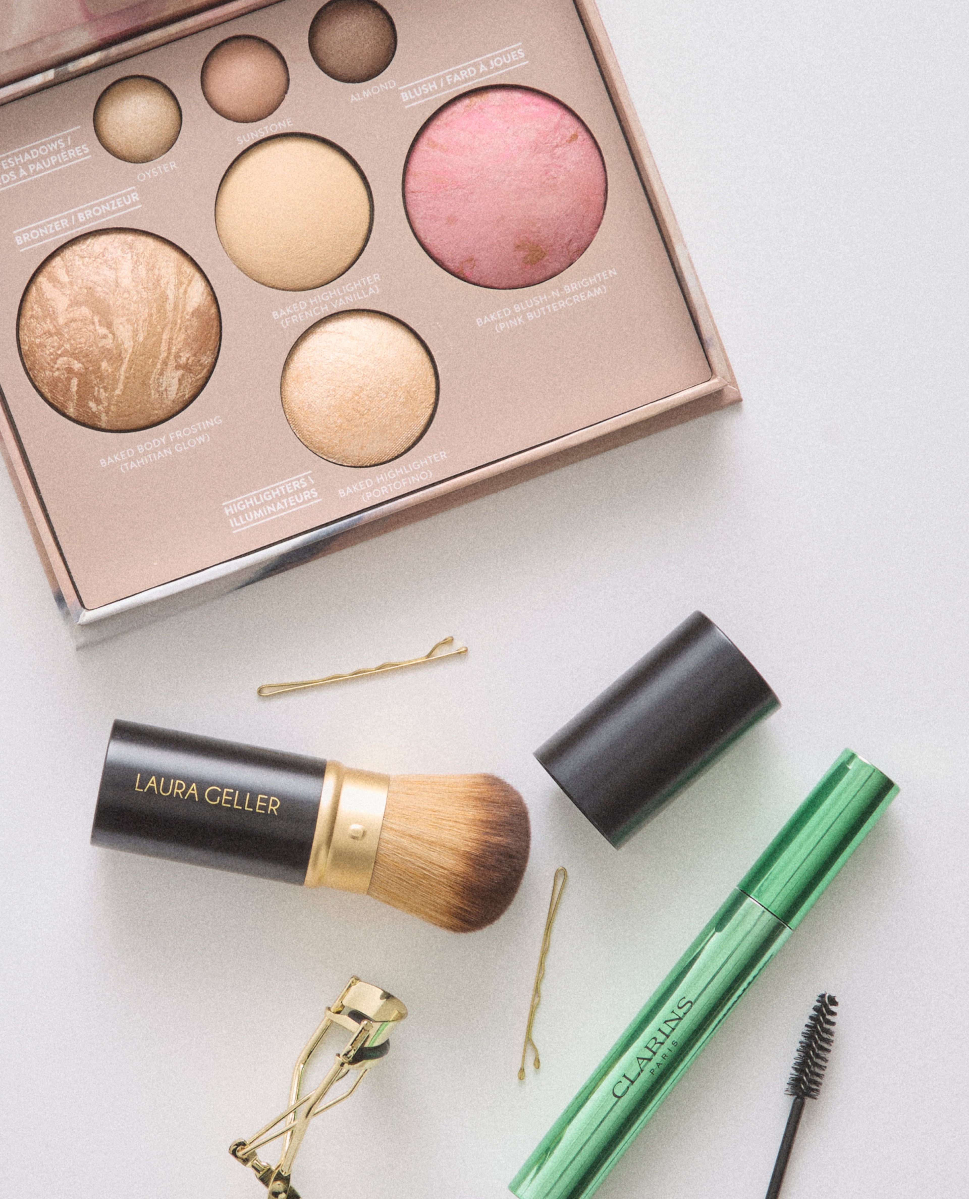 Flat lay of makeup products including a palette, brush, mascara, and eyelash curler on a light surface.