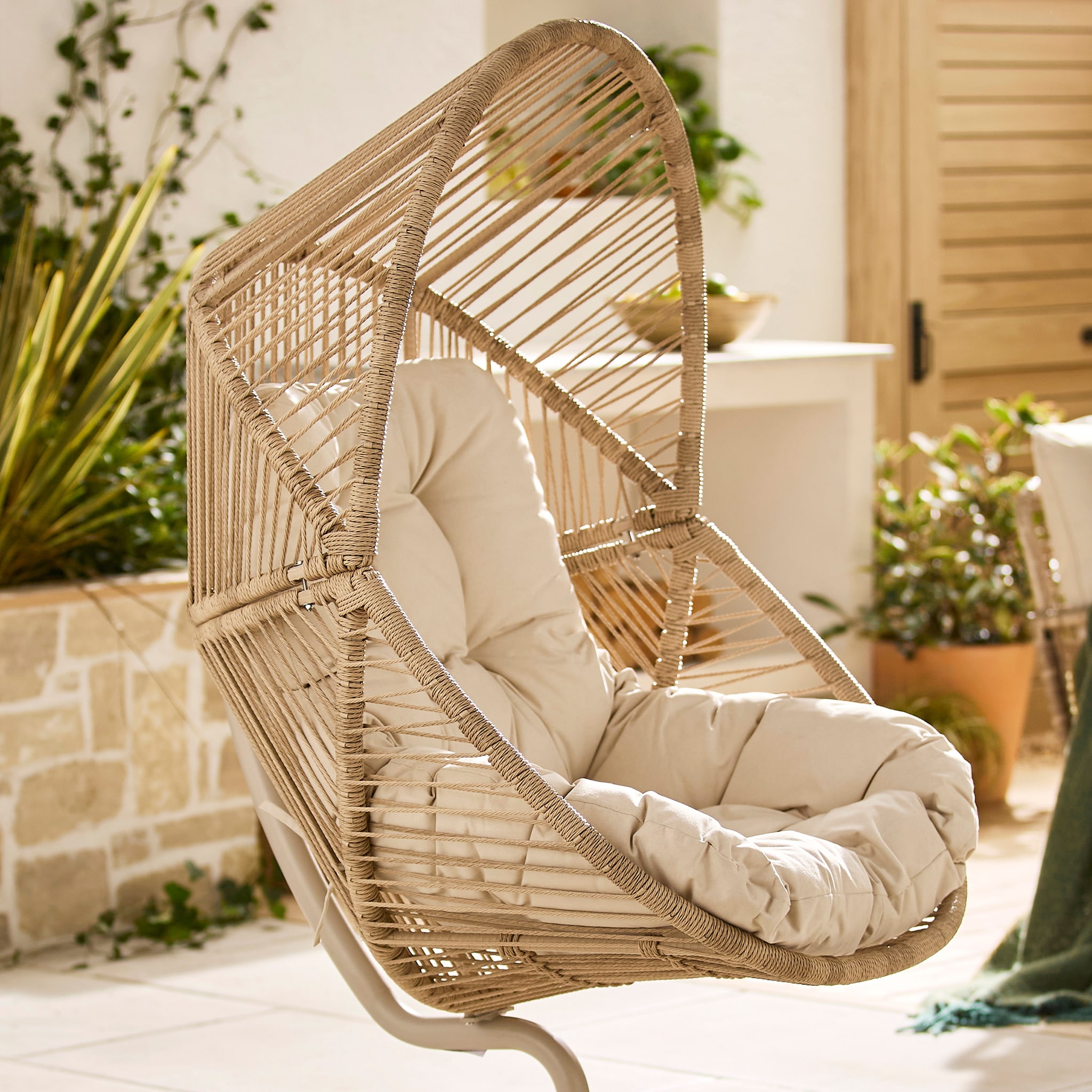 A stylish rattan hanging egg chair with a soft cream cushion, set in a bright, sunlit garden surrounded by lush green plants and natural stone.
