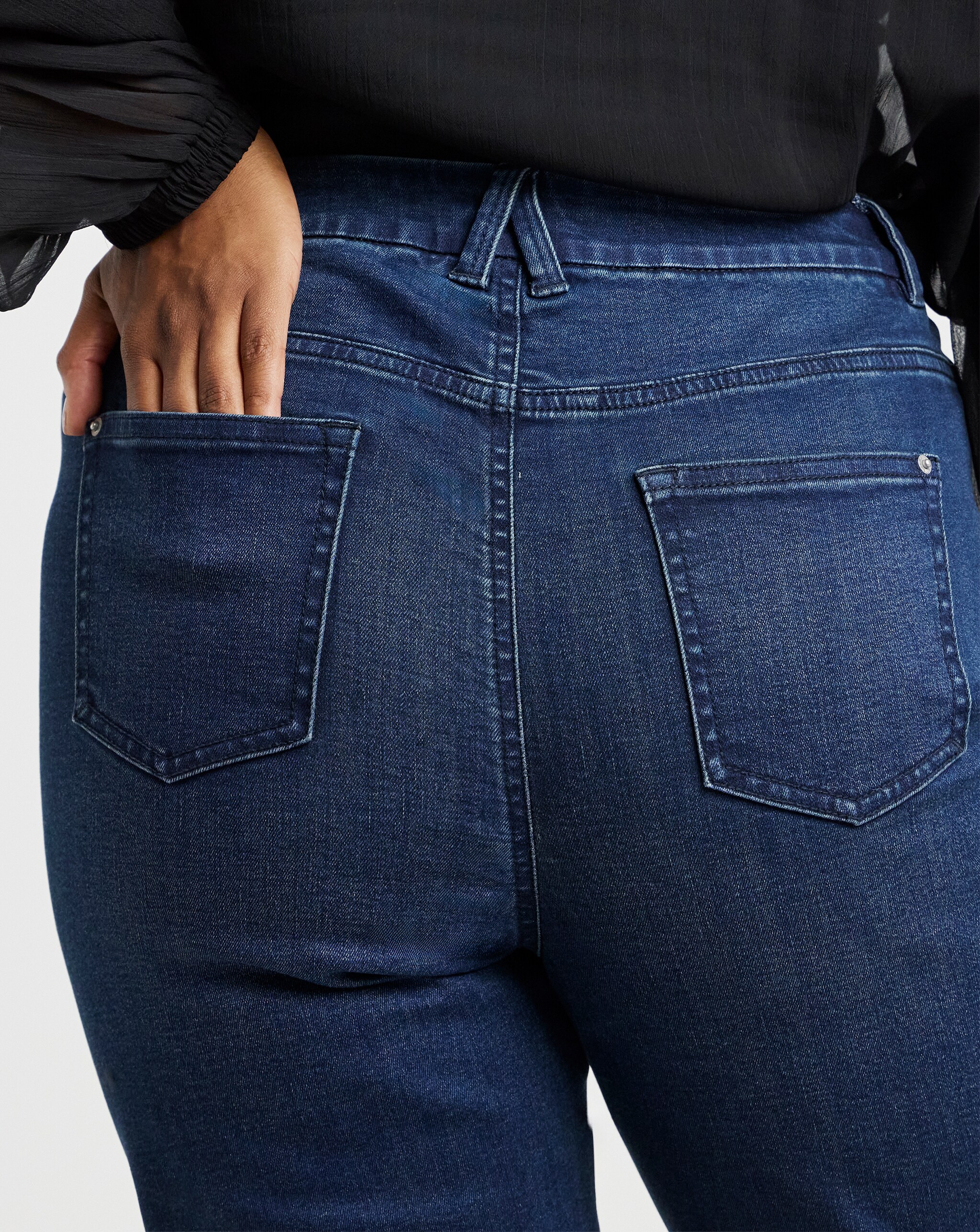 Close-up of dark blue denim jeans from the back, showing two pockets and a hand placed in one pocket.