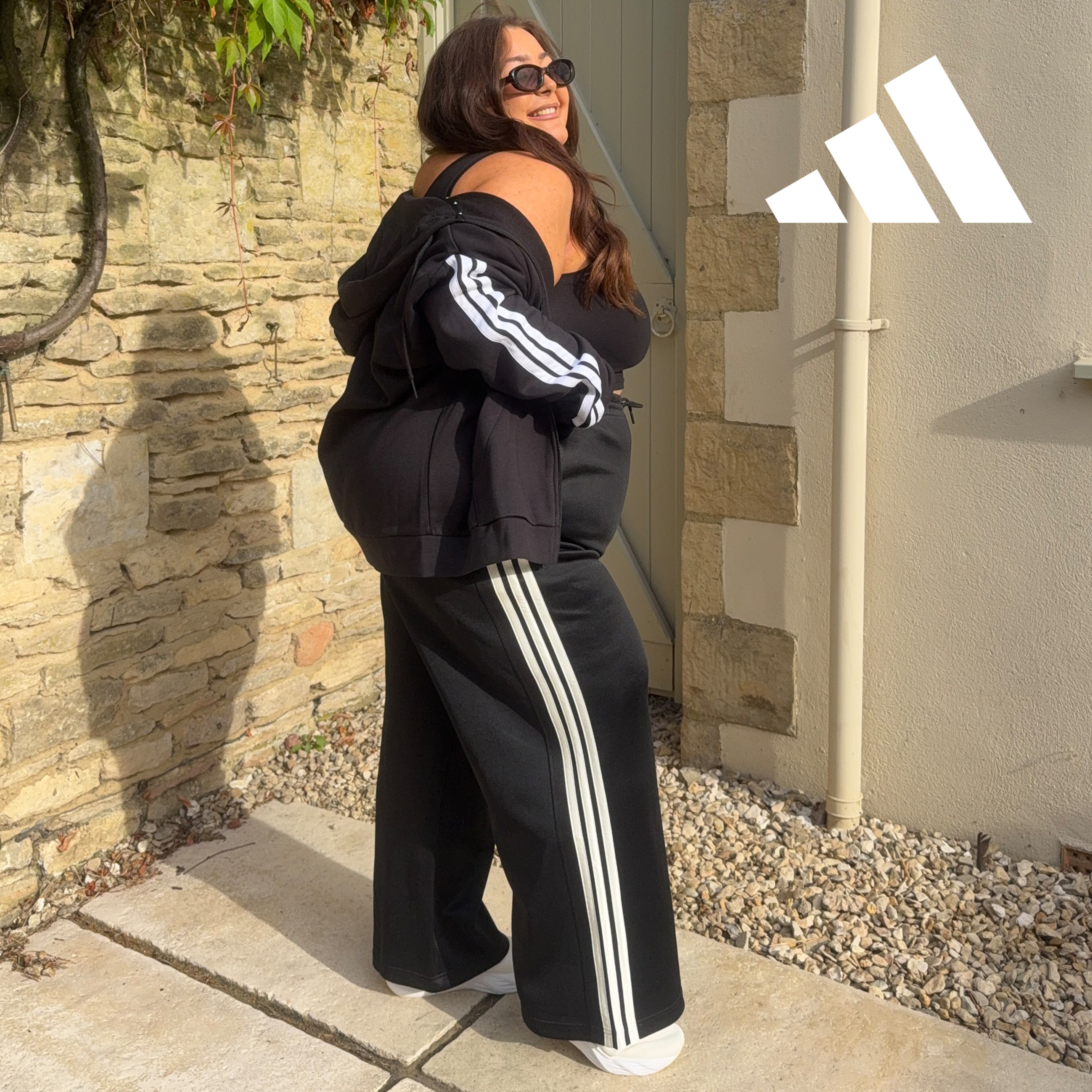 Model wearing adidas black sleeveless fitted top, black wide-leg track pants with three white side stripes, and a matching black zip-up hoodie draped off one shoulder. 