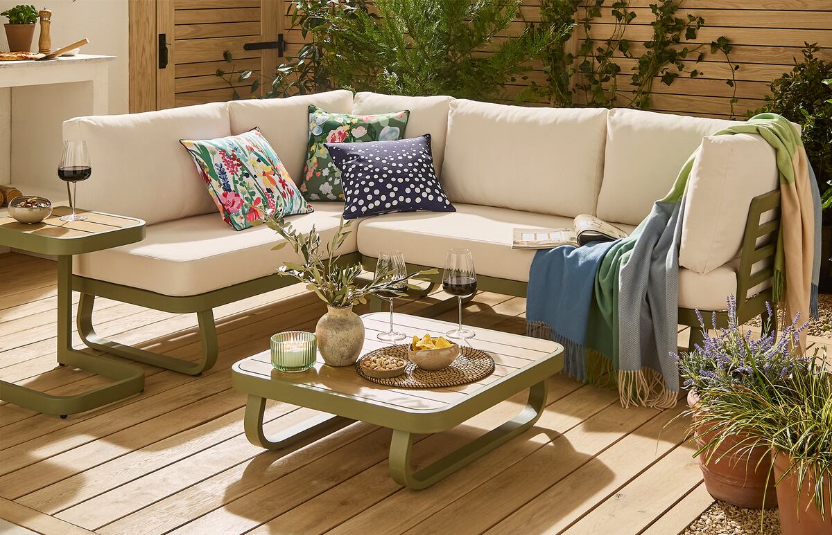 A stylish outdoor seating area featuring a cream corner sofa with colourful floral cushions, green and blue throws, and coordinating olive-green coffee tables. Set on a warm wooden deck, the space is surrounded by lush plants and greenery, creating a bright and inviting garden retreat.