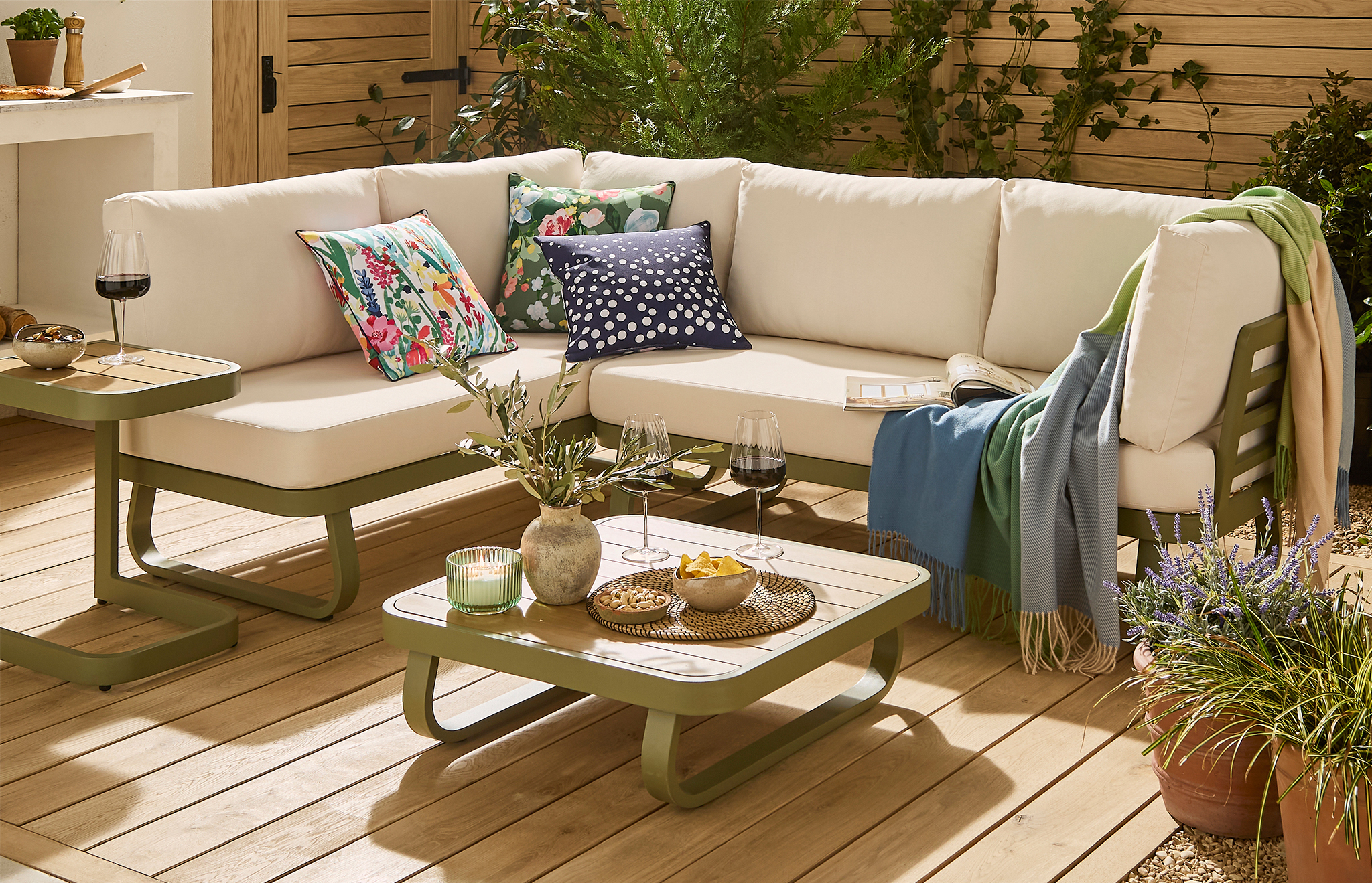 A stylish outdoor seating area featuring a cream corner sofa with colourful floral cushions, green and blue throws, and coordinating olive-green coffee tables. Set on a warm wooden deck, the space is surrounded by lush plants and greenery, creating a bright and inviting garden retreat.