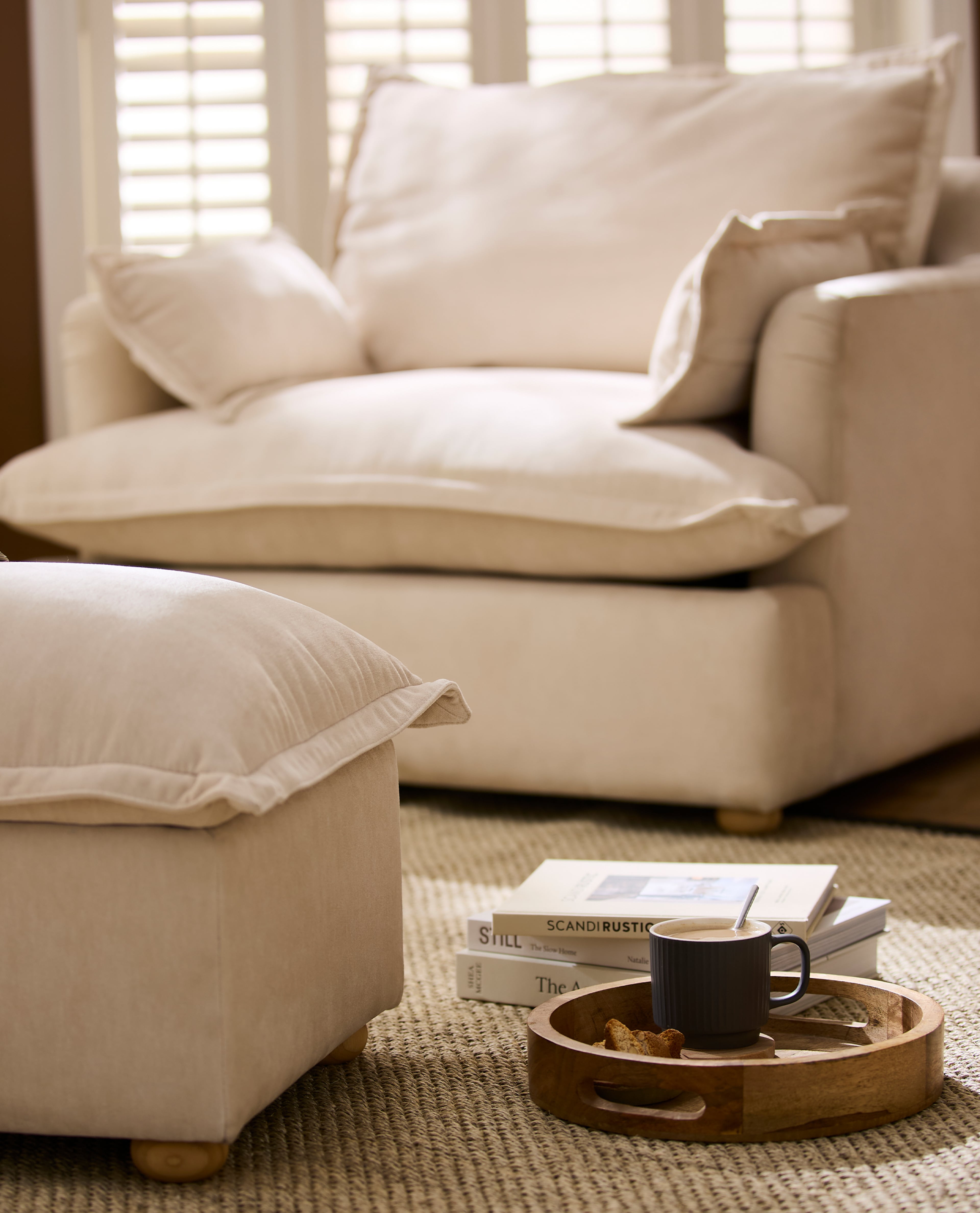 Cream fabric armchair and matching footstool in a cosy living room with a wooden tray, mug and books on a woven rug.