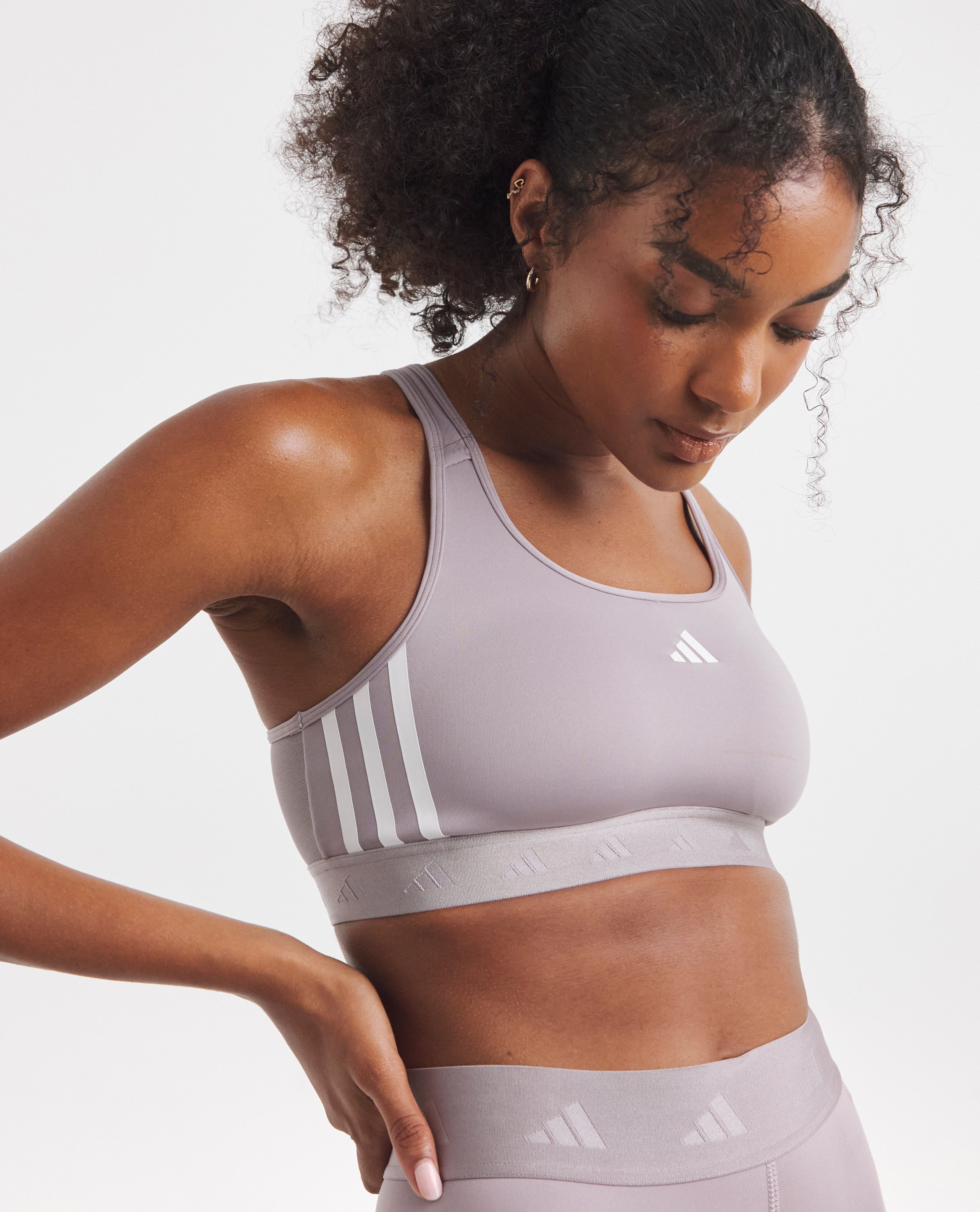 Model wearing a light grey Adidas sports bra and matching leggings, looking down in a studio setting.