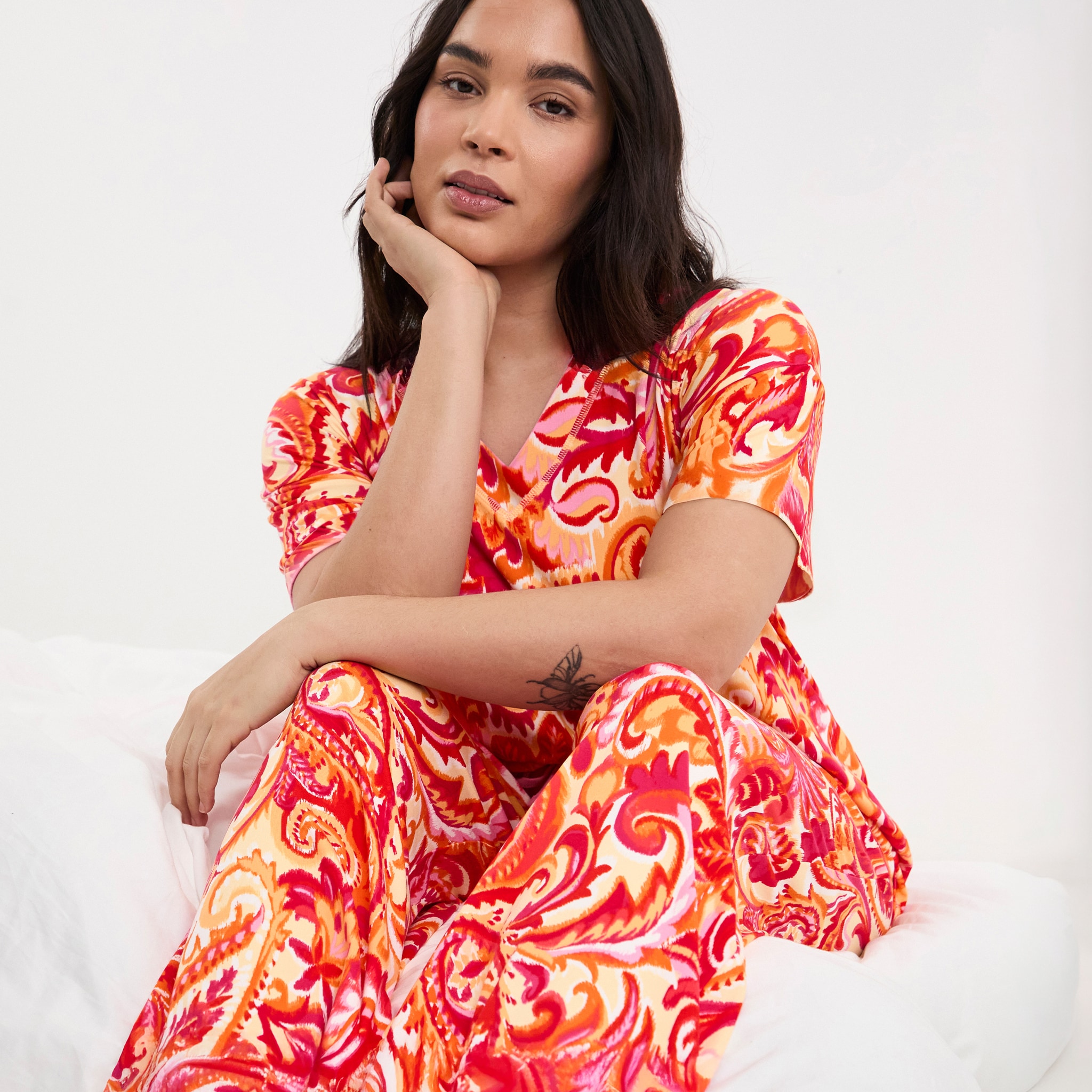 Person seated against a white background wearing a vibrant orange, pink, and cream patterned outfit, resting a hand against their face.