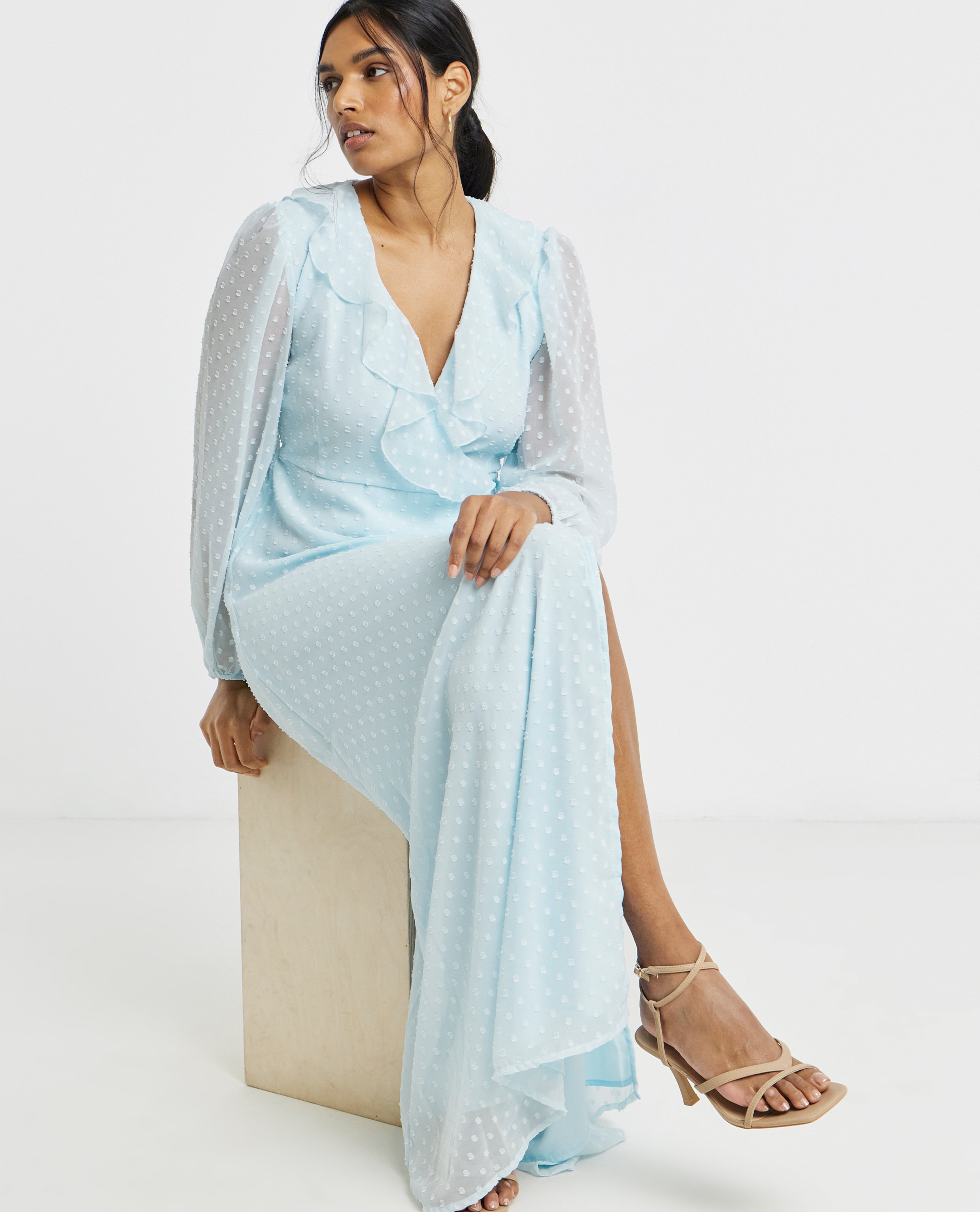 Person seated on a wooden block wearing a light blue long-sleeve maxi dress with ruffle detail and strappy heels against a plain background.