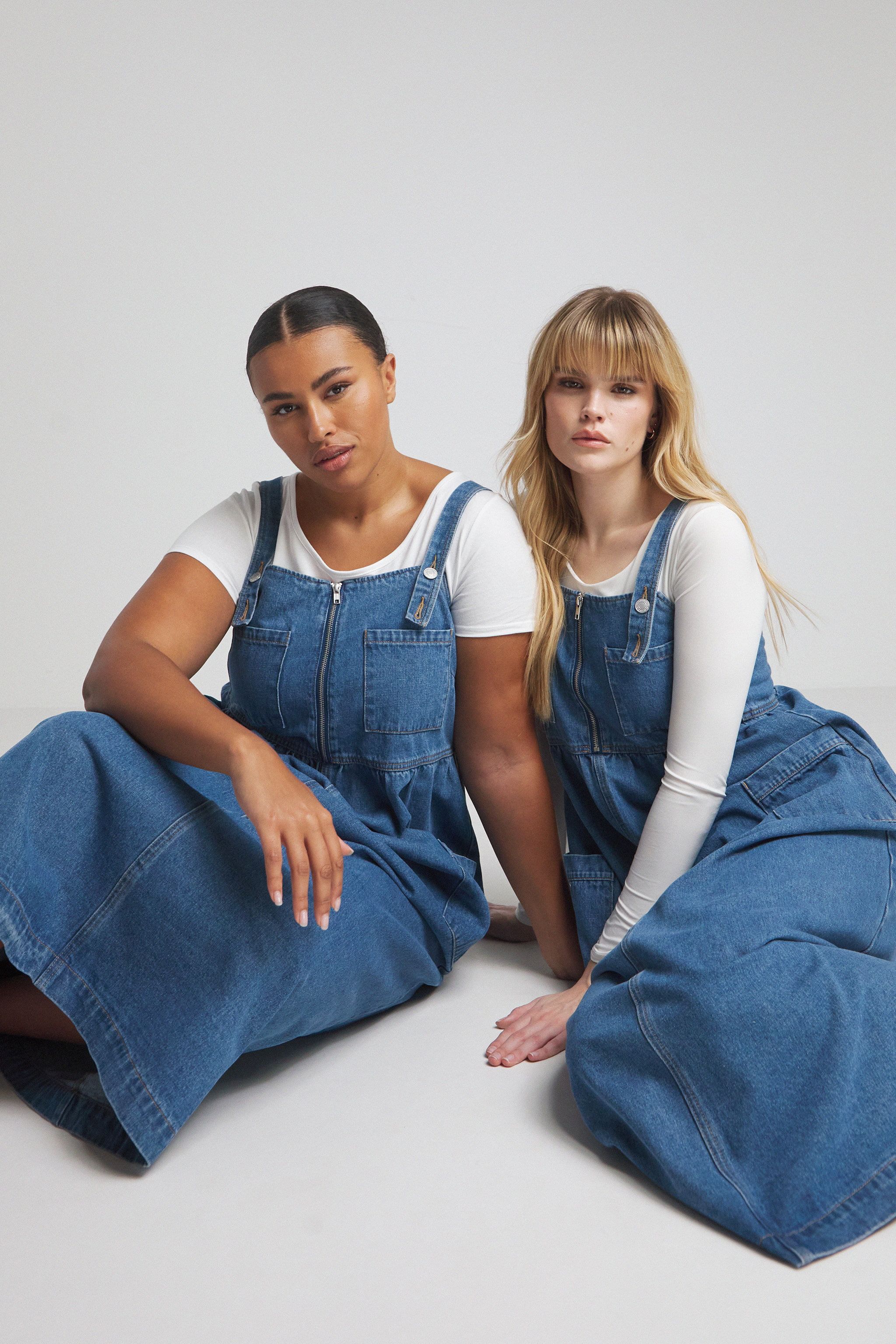 Two models wear the apron denim dress in mid-blue